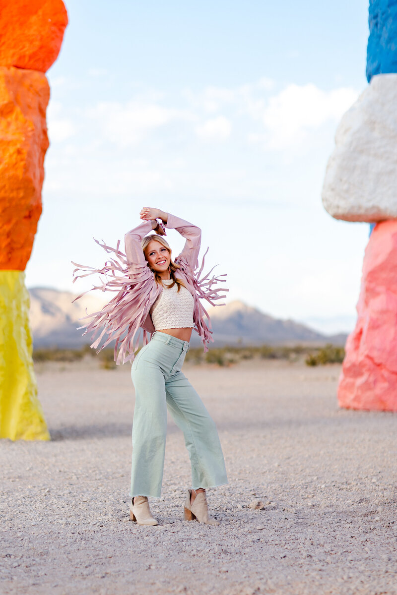 High school senior twirls in the Las Vegas desert wearing a pink fringe jacket and pastel green pants, her outfit flaring in motion as she dances with joy at Seven Magic Mountain during a Brooke Tucker senior session