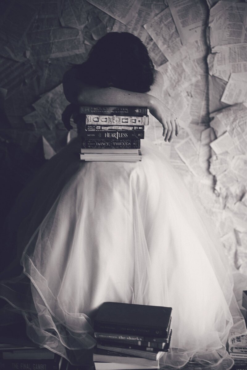 A woman in a white dress is sitting on top of a stack of books, captured by Shreveport senior photographer Britt Elizabeth.