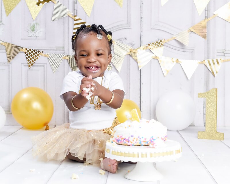 A smiling baby girl clapping her hands in a gold tutu with pearl accessories, sitting with gold balloons and bunting in a glamorous cake smash setup.