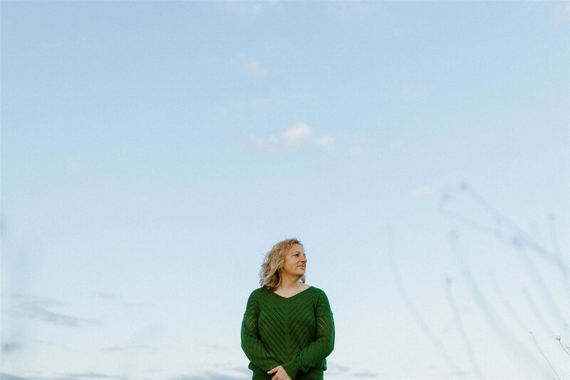 Writer, Sarah Freymuth, stands among trees in a blue t-shirt smiling and looking into the distance