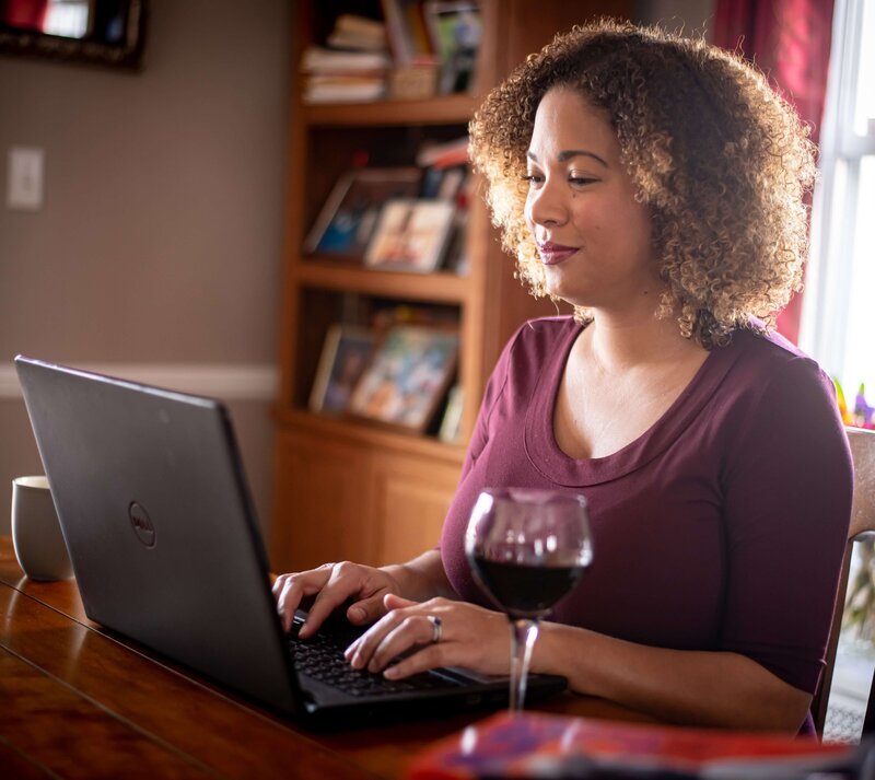 woman sitting at a dining room table typing on a computer with wine
