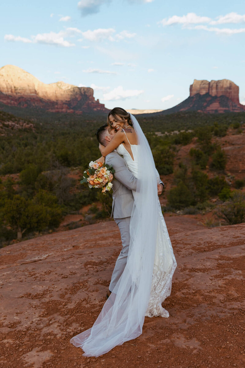 Groom lifts smiling bride with bouquet at Sedona’s Cathedral Rock trail.