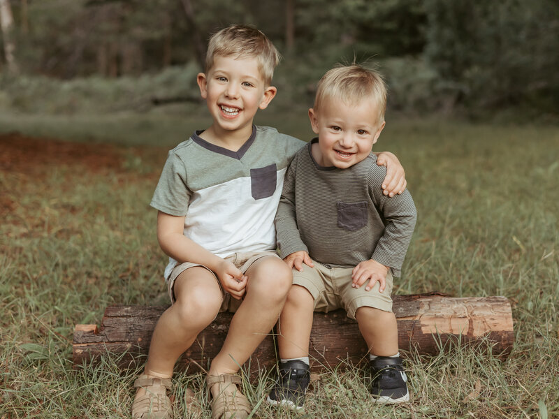 Big and little brother sitting on a log together smiling for their family photography session.
