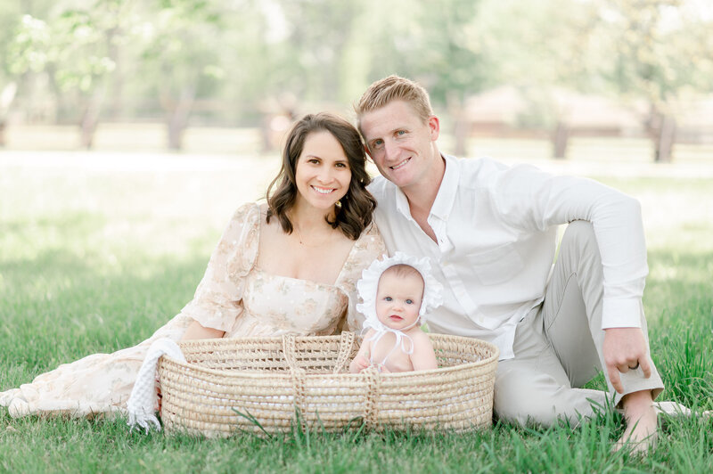 A young nashville family smiles with their baby girl sitting in a moses basket by luxury newborn photographer kristie lloyd