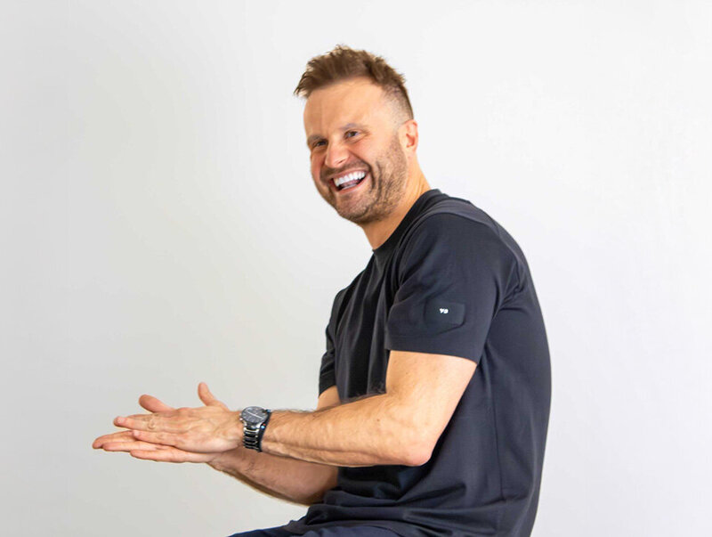 Smiling man in a black t-shirt clapping joyfully in front of a plain white wall, posed casually for a profile-style photo.
