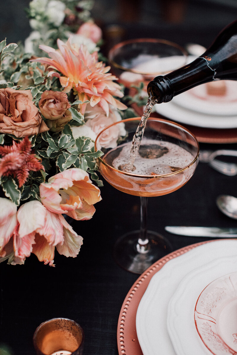 Champagne is being poured into a peach coupe glass which sits on a black wedding reception table set with copper chargers, vintage depression glass plates, mercury glass candles and peach and copper flowers