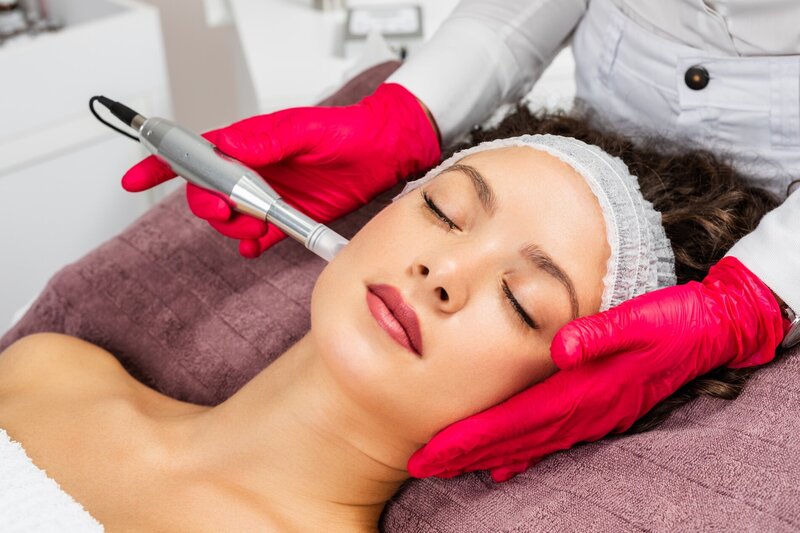 Closeup image of a woman lying down getting microneedling treatment from an esthetician with hot pink gloves