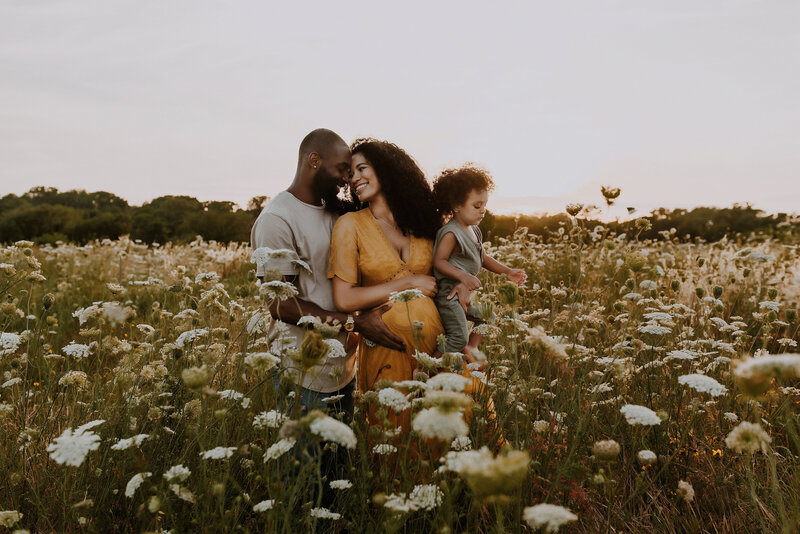 A mom, dad and son standing together very close while dad touches mom's pregnant belly in a field of tall white flowers at White Rock Lake in Dallas, Texas