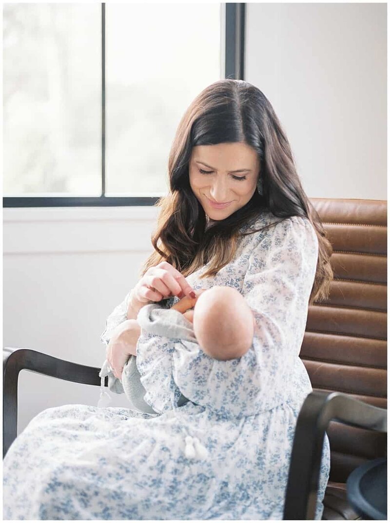 A mother wearing a blue floral dress holds her infant son while sitting in a brown chair photographed by DC Newborn Photographer Marie Elizabeth Photography