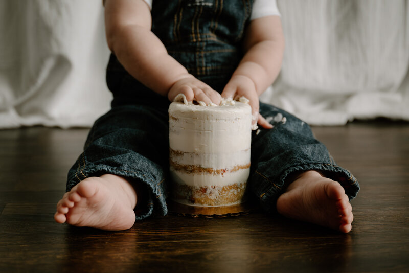 Photo of one year old boy in jean overalls pushing hands into a small white cake in Baltimore Maryland.