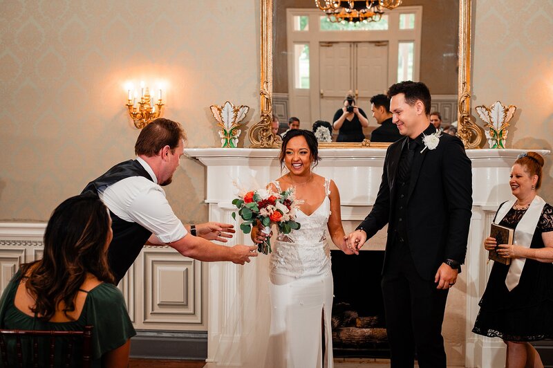 The father of the bride, wearing a black vest and white shirt, leans in to hand the bride her bouquet at the end of the wedding ceremony. The bouquet is crimson, orange and ivory. The bride is wearing a fitted wedding dress with a thigh high slit and spaghetti straps. The groom is wearing a black suit with a white boutonniere. The officiant, dressed in black with a white sash, stands off to the side at Oaklands Mansion in Maney Hall.