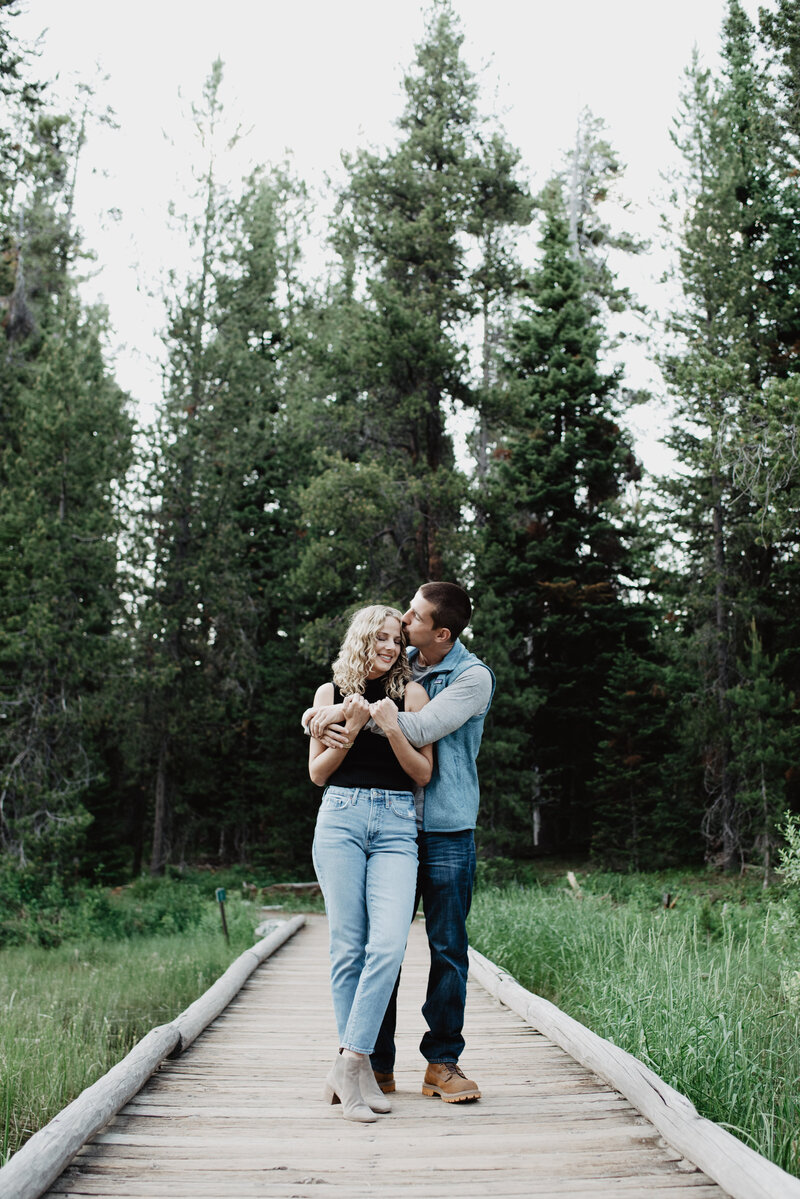 engagement photos on a bridge in the Tetons with tall pines behind the couple as the man wraps his arms around his bride and kisses her head