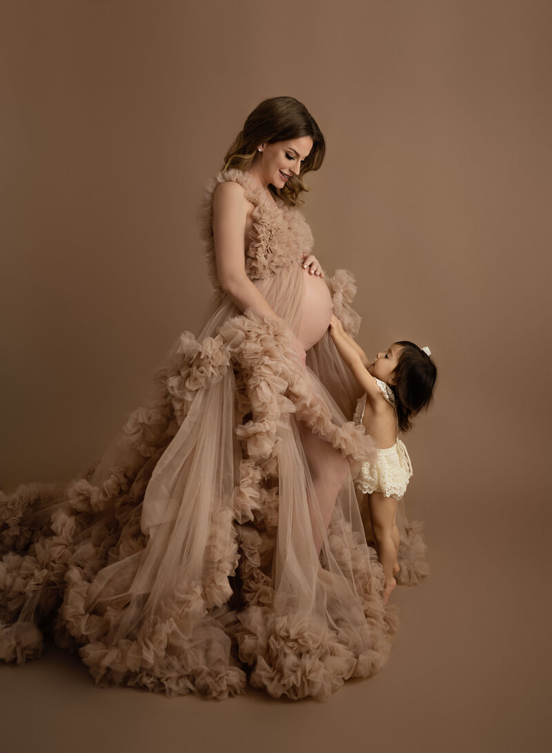 Pregnant women dressed in tan tulle gown with belly out. Toddler is reaching up and touching Mom's belly.