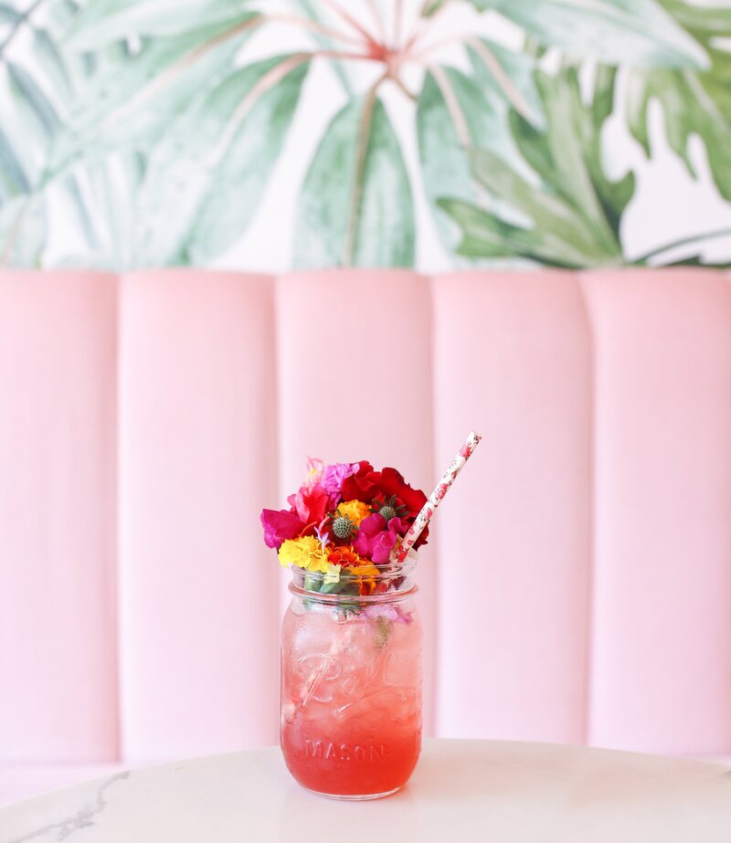 a  cute close up shot of a pink drink served in a mason jar and garnished with a small bouquet of flowers in a pink booth with tropical wallpaper socialfizz