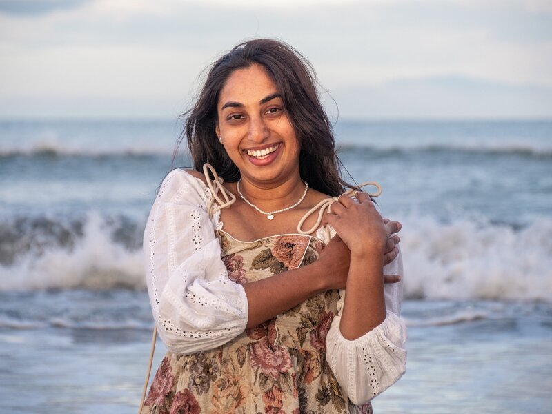 a woman with long dark hair stands on the beach, smiling wide, with waves crashing behind her. she wears a flower print dress with white lace sleeves.