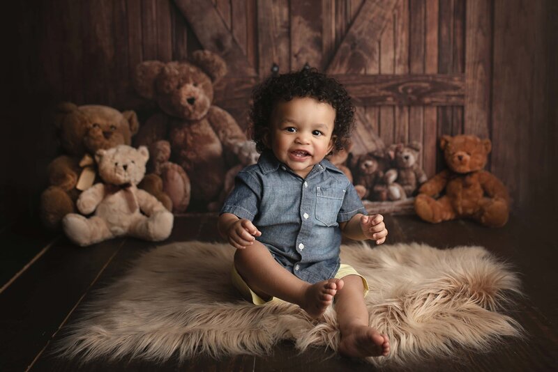 A toddler boy in a denim shirt sits on a fur rug surrounded by teddy bears decorated by a Lafayette Baby Milestone Photographer