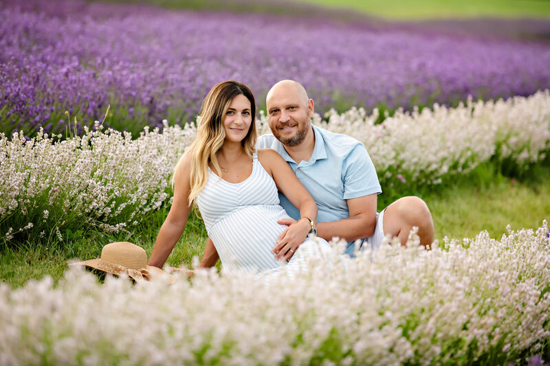 Woman wearing a blue and white dress sitting with her husband on the ground holding her baby belly in a lavender field for her outdoor Burlington Maternity Photography session.