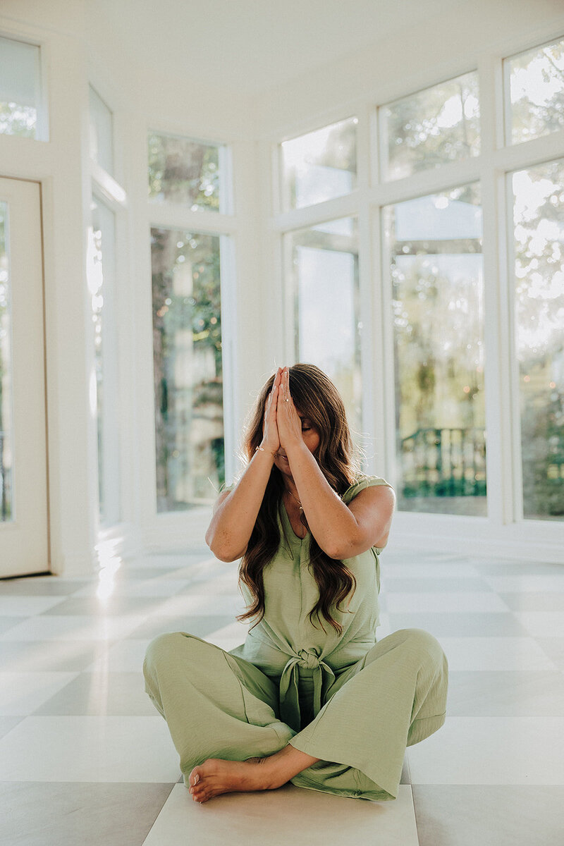 Patty wearing green sitting cross legged on the floor with her hands in prayer at her crown chakra