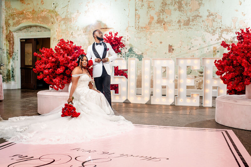 This fashionable Black couple poses amidst bold red florals and oversized marquee letters of their last name at 701 Whaley, with stunning decor by Opulence Events. A perfect blend of modern luxury and timeless romance.