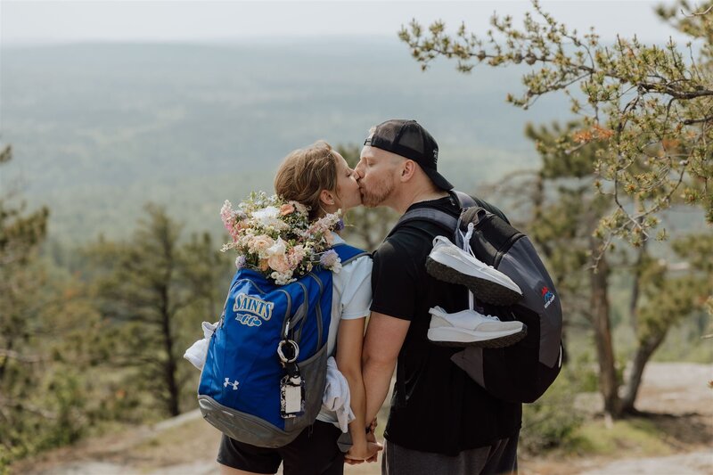 couple in hiking attire kissing and wearing backpacks with a wedding bouquet sticking out of one.