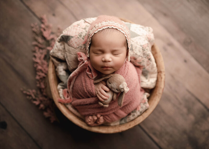 Aerial image. Baby girl is captured sleeping in a wooden bowl for her newborn photoshoot in Murrieta, CA. Baby girl's hands and toes are peeking out of the swaddle. In her hands, she is holding a tiny felt bunny. Captured by best Murrieta newborn photographer Bonny Lynn Reed.