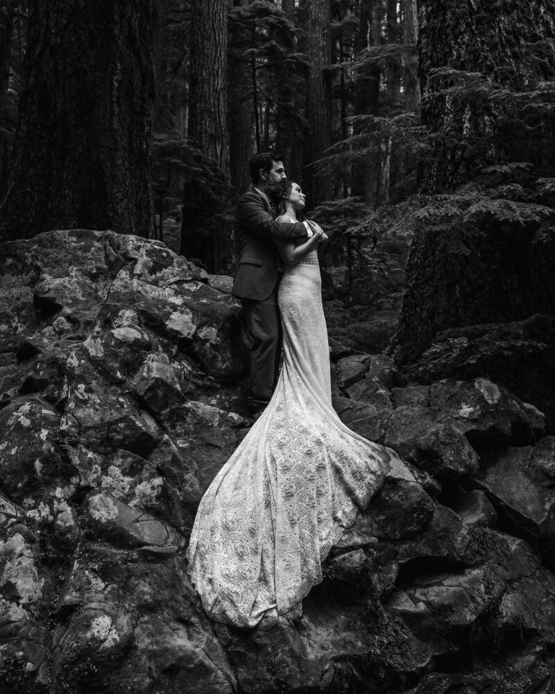 A couple in wedding attire embrace on top of a jagged boulder in the heart of the forest during their olympic national park wedding