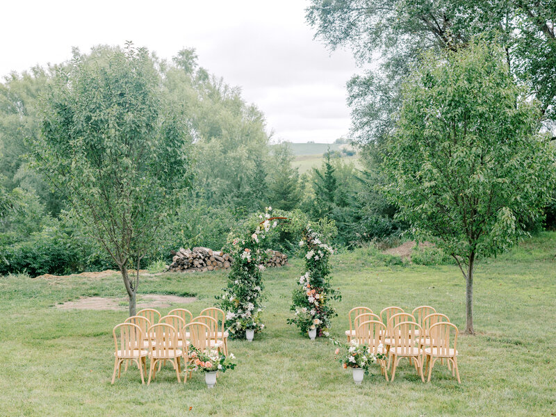Wedding ceremony decor with green, pink, and white floral arch, potted vases, and wooden chairs overlooking a green vista