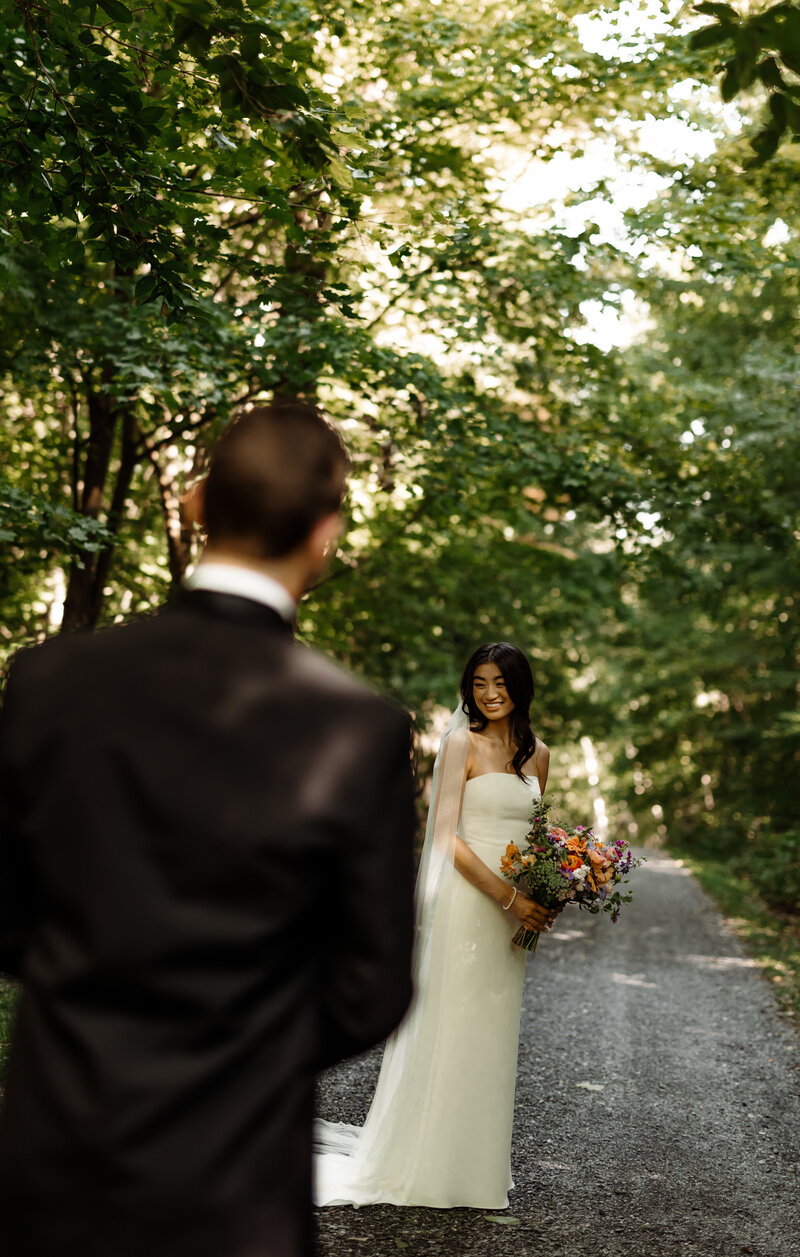 A bride in a white dress stands holding a colorful bouquet on a tree-lined path, smiling at a groom in a black suit whose back is to the camera. Sunlight filters through the leaves, casting a warm glow, capturing the essence of fine art weddings and portraits in New England.
