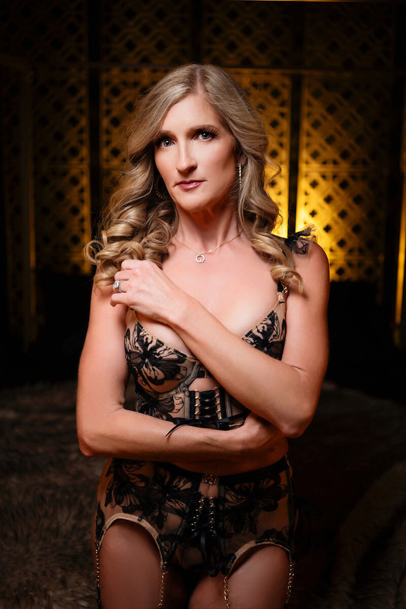 A woman with long, wavy hair poses confidently in floral-patterned lingerie. The background is dimly lit with a warm, intimate glow.