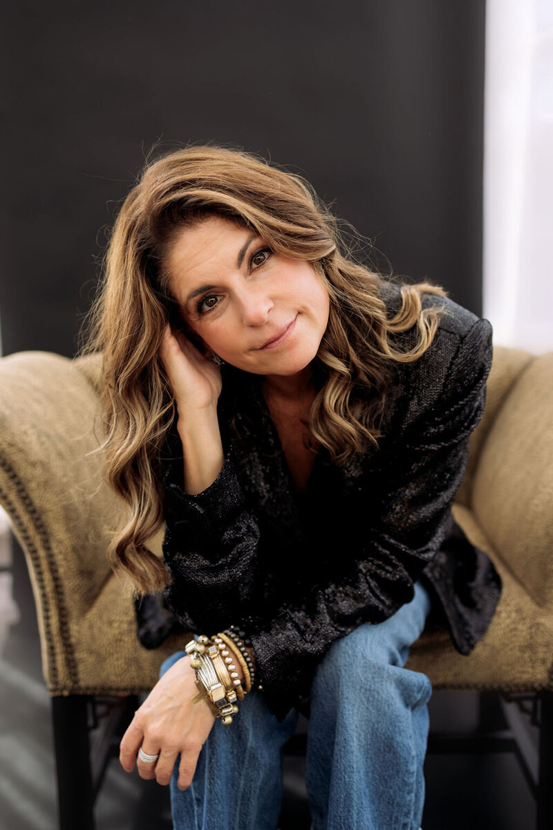 Kerri Link sits on a soft armchair, smiling warmly and inviting connection through healing, creativity, and purpose.