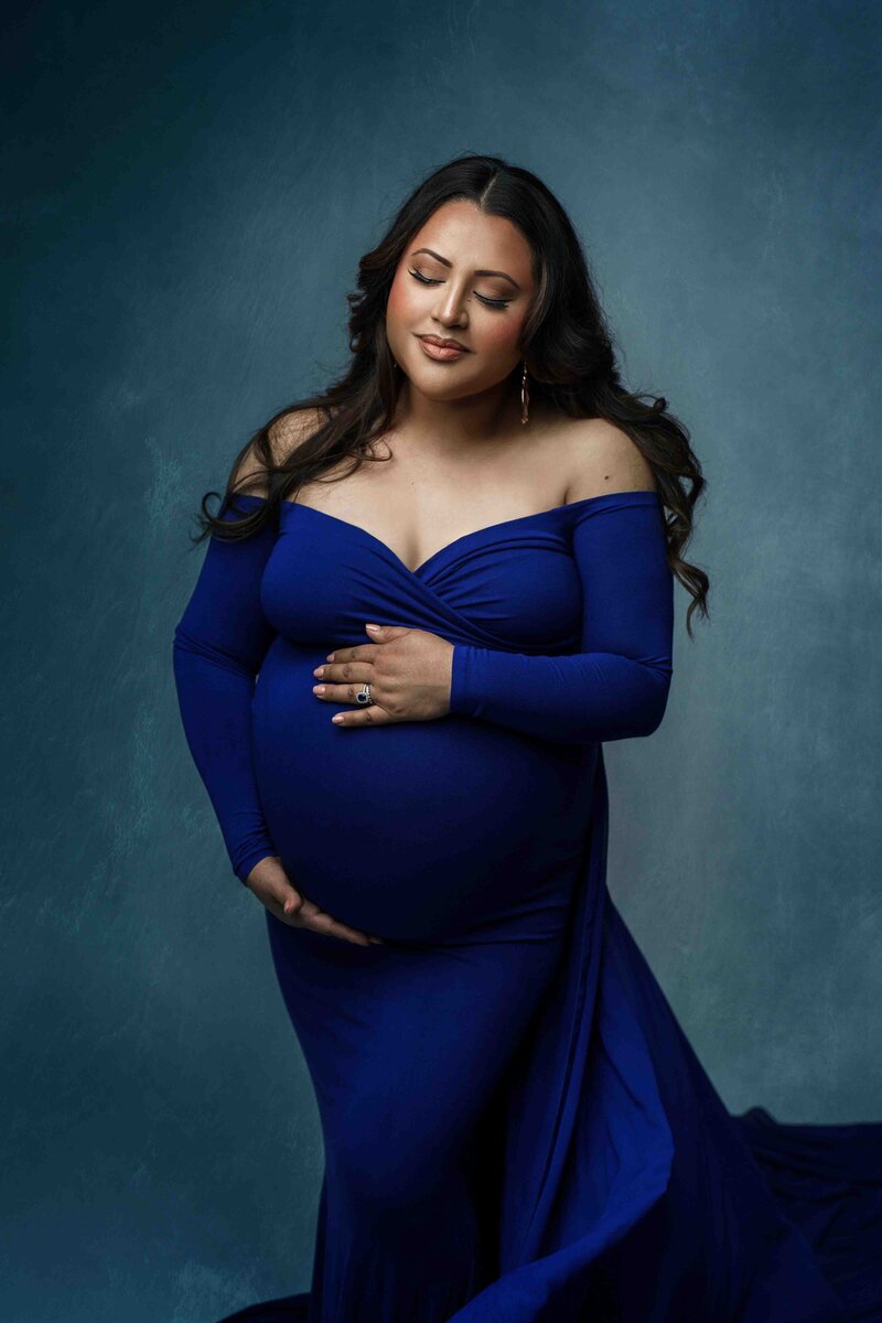 Tacoma mother-to-be poses in studio with hands embracing her belly while wearing a royal blue dress. She's smiling with eyes closed.