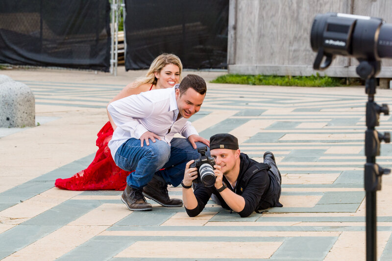 Travis Stitt lying on the ground while showing an engaged couple a preview of their photo on the back of his camera during an engagement session, highlighting his unique perspective and dedication to capturing perfect moments.