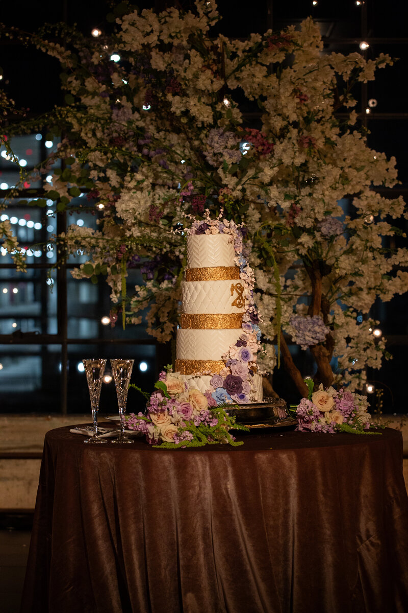 Mini foldable round table covered in dark satin linen holding a tall wedding cake with gold bands and purple florals, styled against a moody indoor reception backdrop.