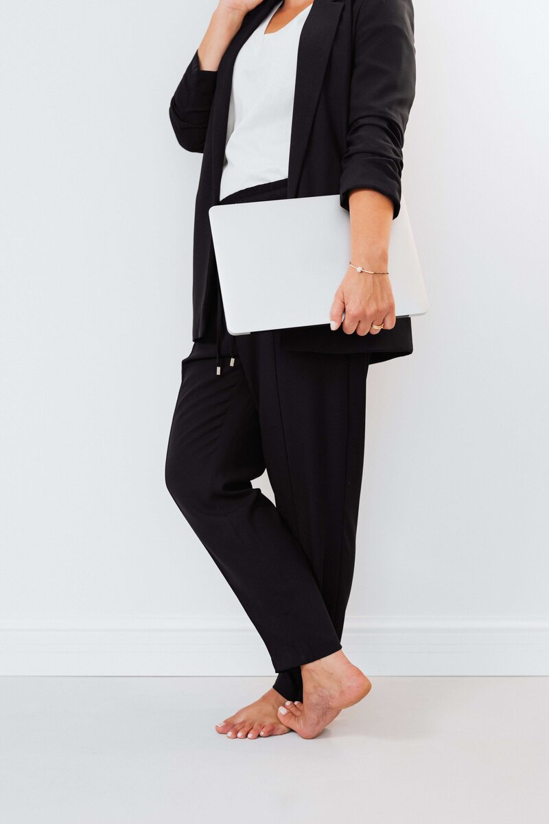faceless barefoot woman wearing all black in a white room holding silver laptop