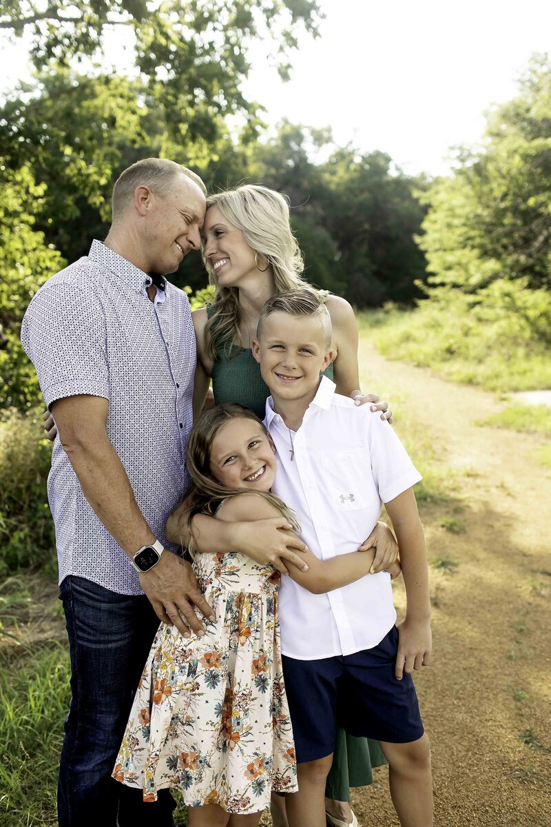 Smiling family hugging tightly while sitting on the grass at Northwest Community Park, with trees and greenery in the background