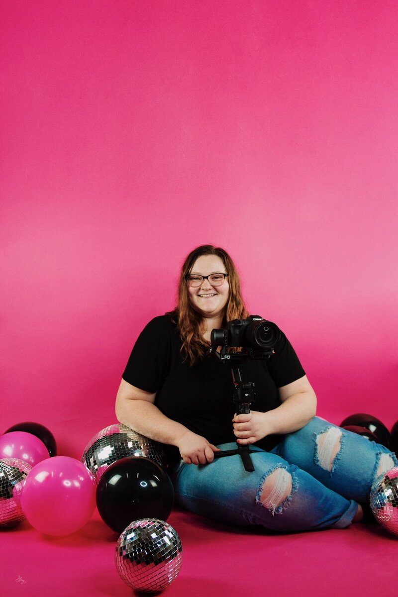 portrait of mara shields, owner of M20 Media, sitting and smiling while holding her camera and gimbal, with balloons, disco balls, and a hot pink background