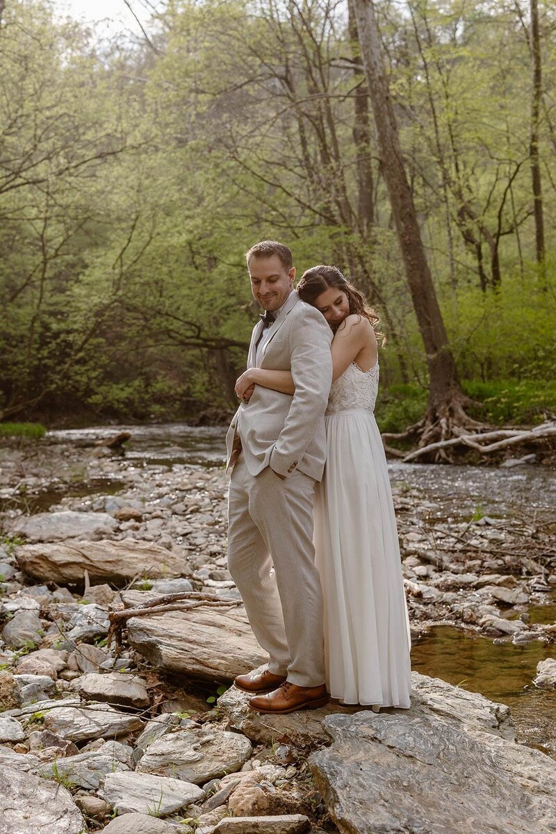 A bride and groom in the woods on their wedding day. They are standing next to one another. The groom is kissing the brides forehead, and the bride is smiling, and holding up her dress