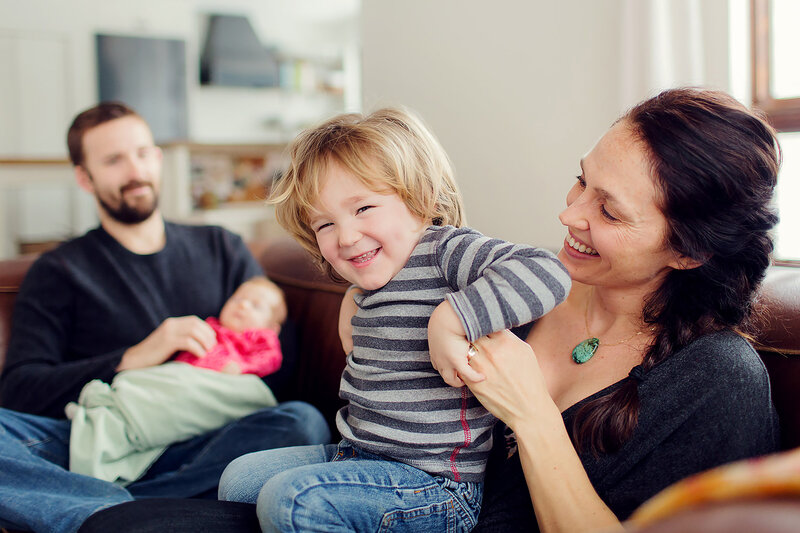 At-home shoot with mom and toddler on couch laughing, Dad is holding newborn sister behind them.