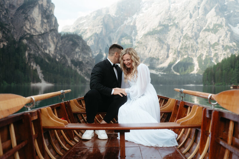 Dolomites Elopement | Groom whispers to bride as she laughs in a wooden boat with Lago di Braies in the background