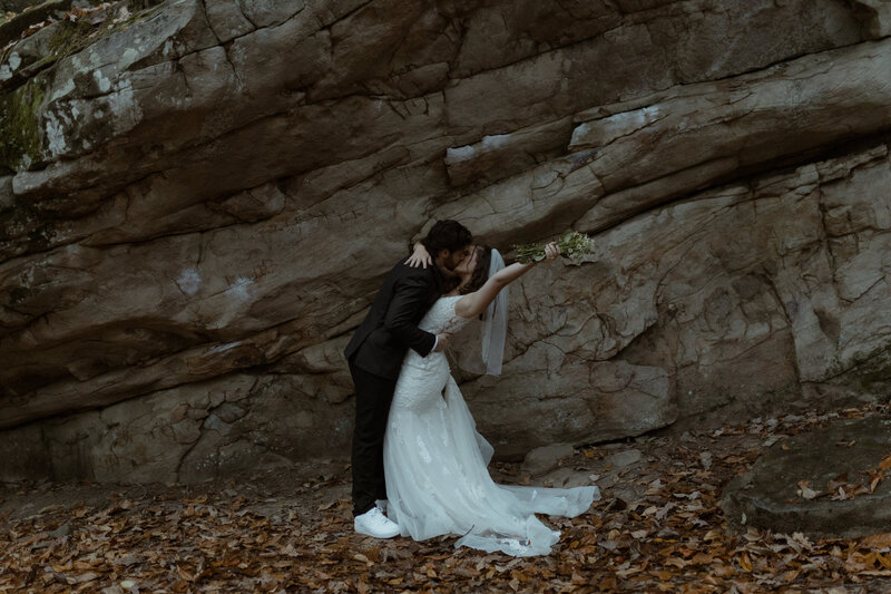 couple sharing a passionate kiss in front of a rock wall, surrounded by fallen autumn leaves. The bride, dressed in a lace wedding gown and veil, leans into a dramatic dip with her bouquet raised joyfully in one hand. The groom, in a dark suit and white sneakers, holds her securely as they embrace.