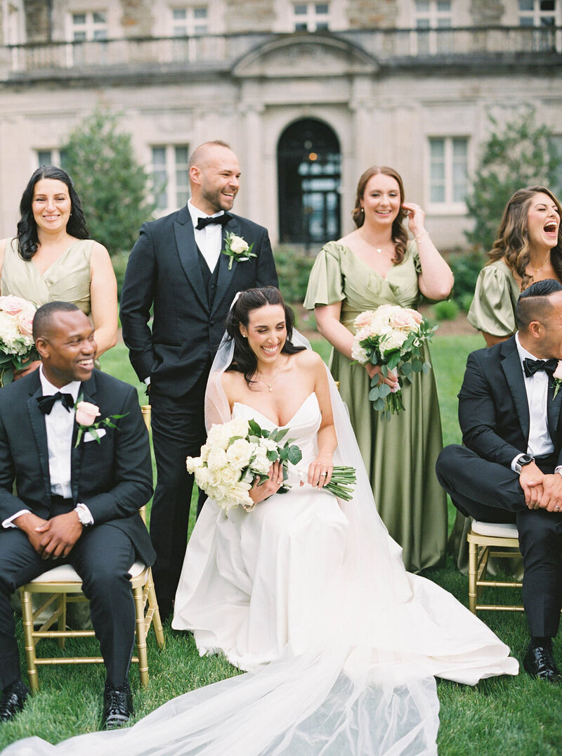 Newport Wedding Photographer | A bride in a white gown sits smiling with bridesmaids in green dresses and groomsmen in black suits, posing in front of a historic building. Everyone looks joyful.