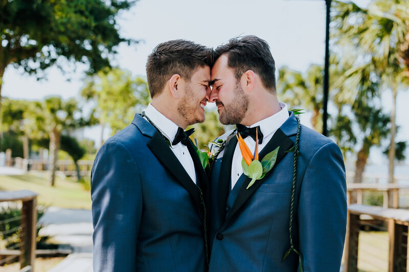 Two grooms in wedding attire embrace at a tropical wedding venue