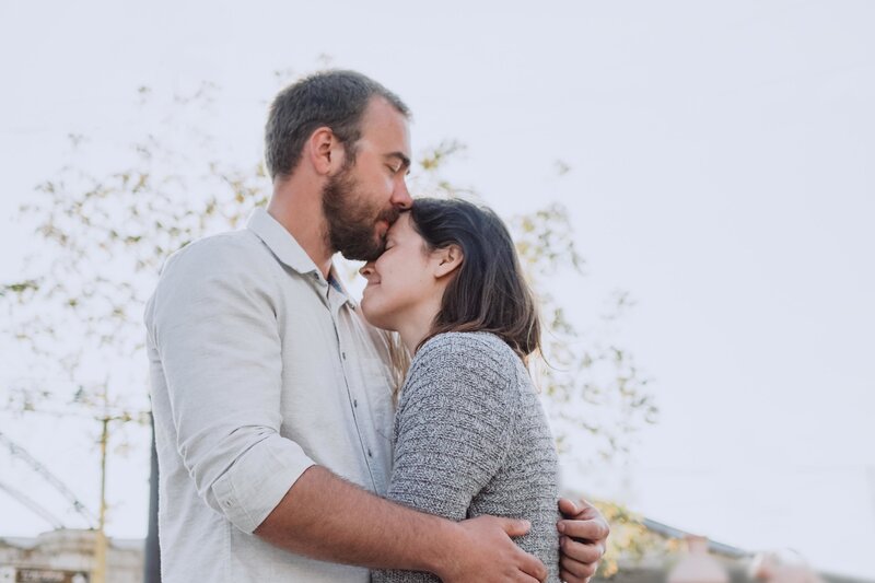 A man kisses his partner on the forehead as he wraps his arms around her. This could symbolize the strength of a relationship after infidelity recovery. Contact a marriage counselor in Florida for support with your relationship. A couples therapist would love to support you!
