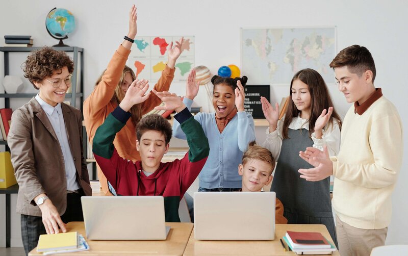 two kids sitting at a desk with laptops hands in the air excited with a group of kids behind them