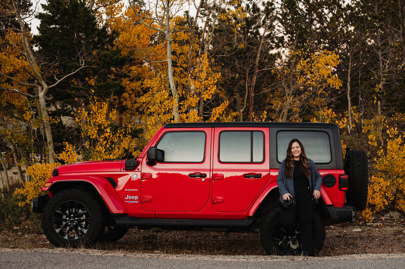 there's a woman sitting on a red jeep wrangler with her hand in the air. She's wearing sunglasses and a hat. There are fall colors in the trees and she's parked on a dirt road. 