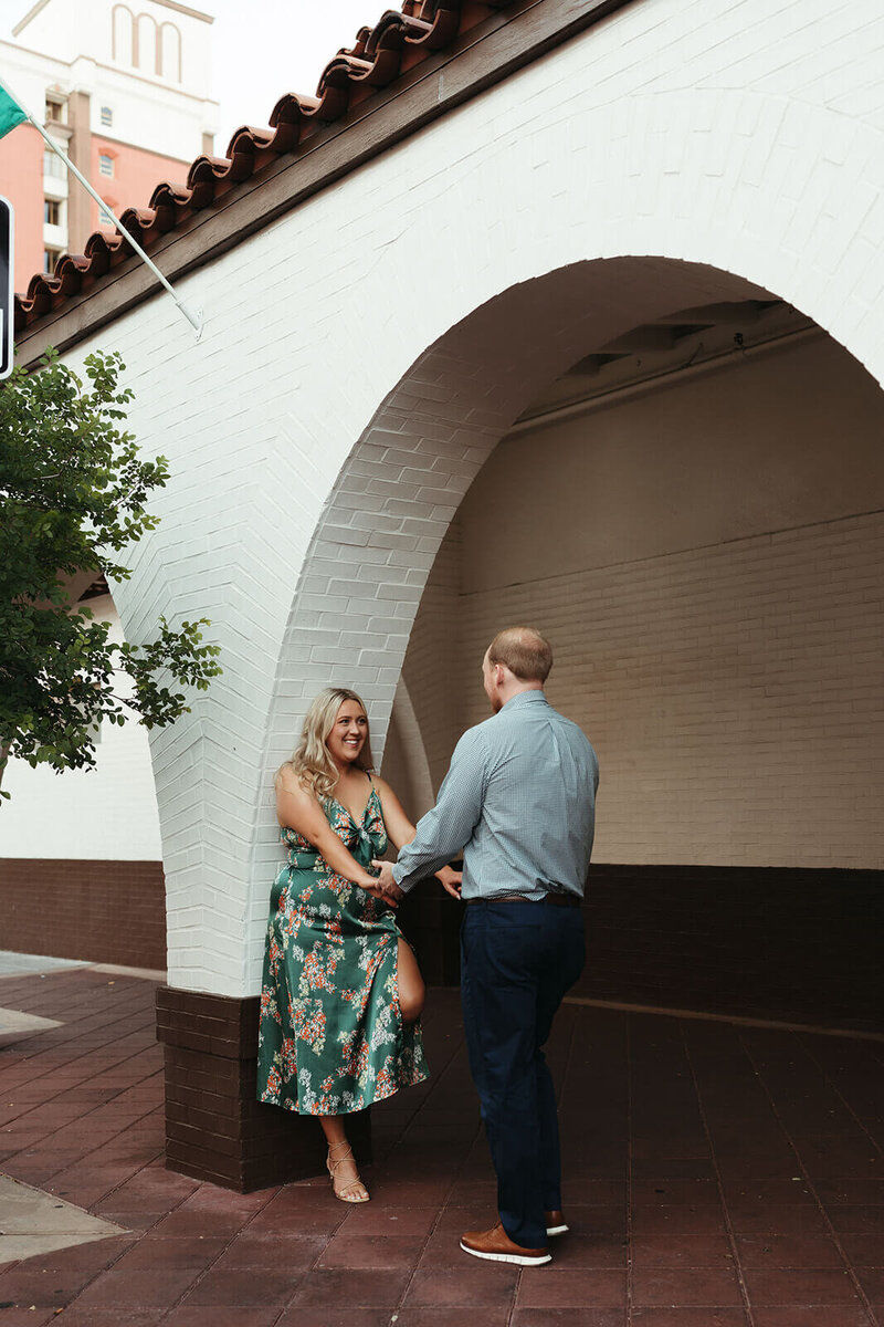 The girl is standing, leaning against the wall, while the guy stands opposite her. They are looking at each other and holding hands