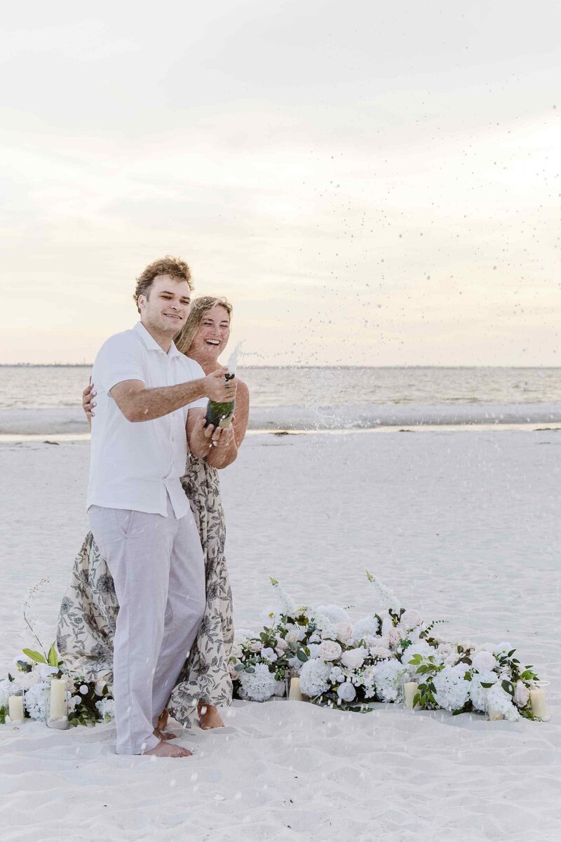 Couple standing next to a flower display, laughing while spraying champagne towards the camera