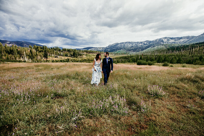 A bride and groom in formal attire stroll together through a wildflower-filled meadow in Lake Tahoe, surrounded by expansive mountain views and dramatic cloudy skies