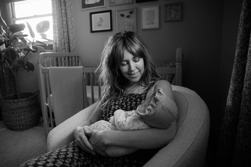 Mother lovingly gazes down while holding newborn in natural light.  