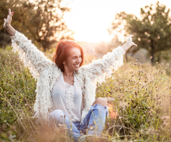 woman sitting cross legged in a field with arms up over her head smiling